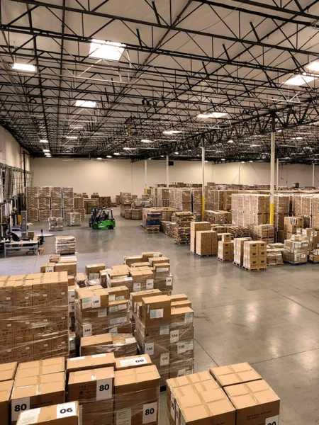 Warehouse interior with palletized inventory, shipping boxes, and forklift operations in a large distribution facility.